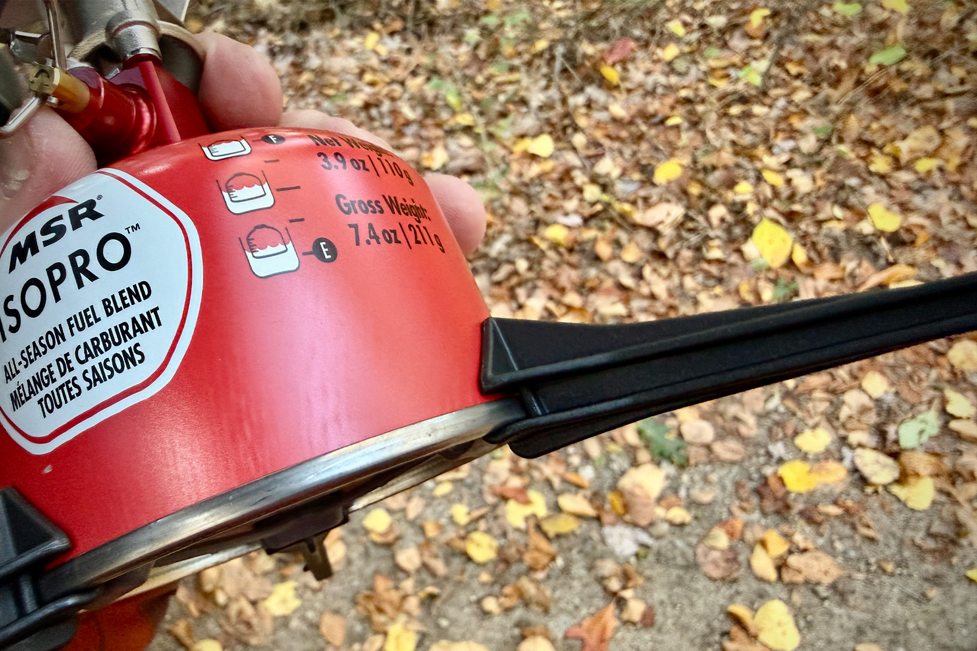 A close-up of a fuel canister with a tripod stabilizer stand on a bed of autumn leaves.