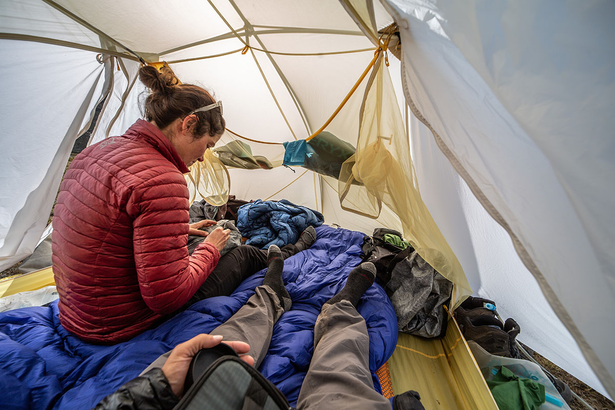 A woman sitting inside a tent with the mesh inner wall visible, looking out at a mountain landscape.