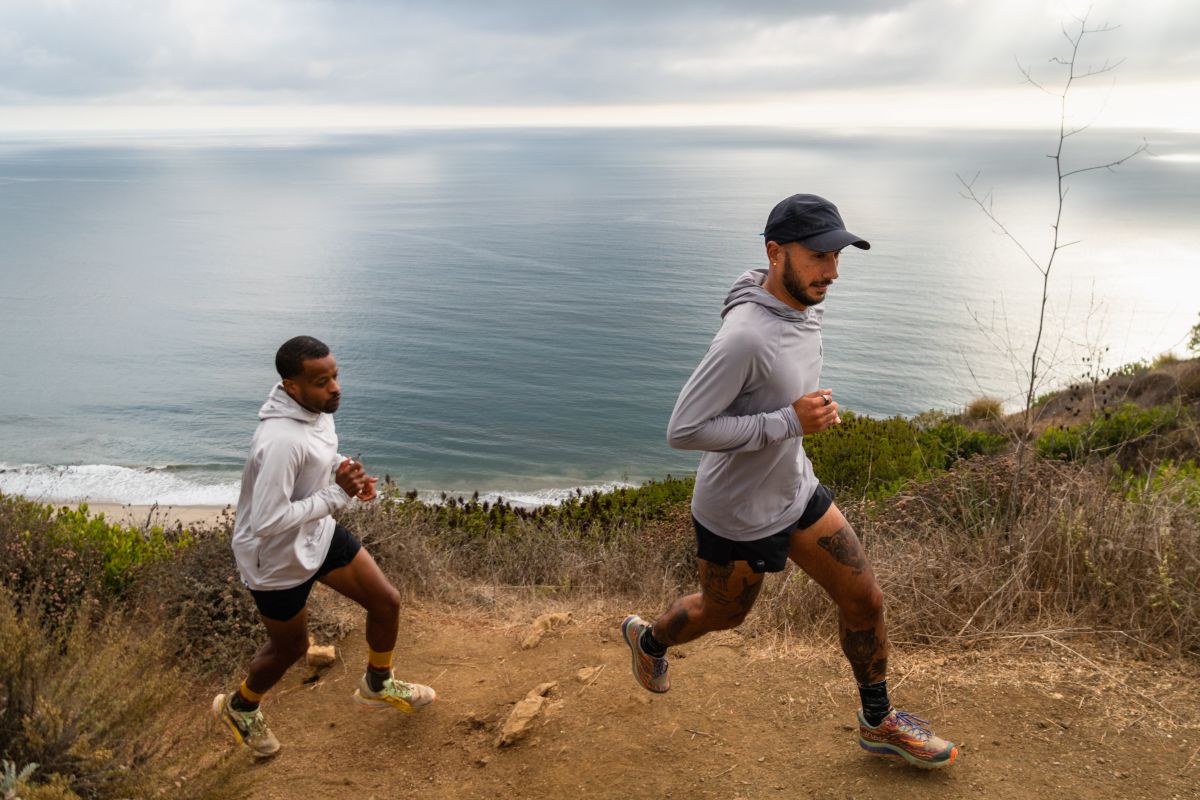 Two runners wearing long-sleeve sun shirts climbing a coastal trail hill.