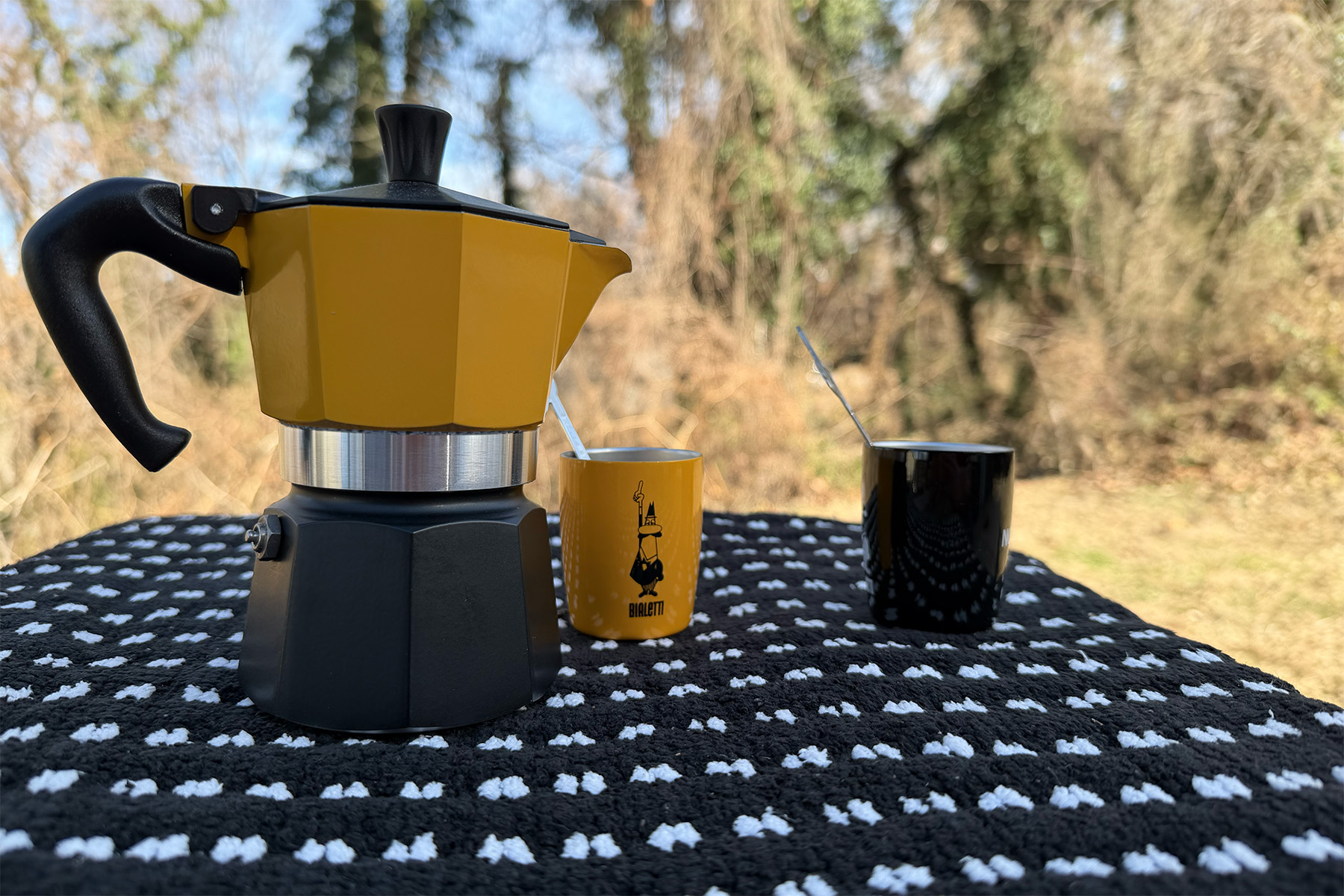 A classic metal Moka pot sitting on a camp table with a scenic forest background.