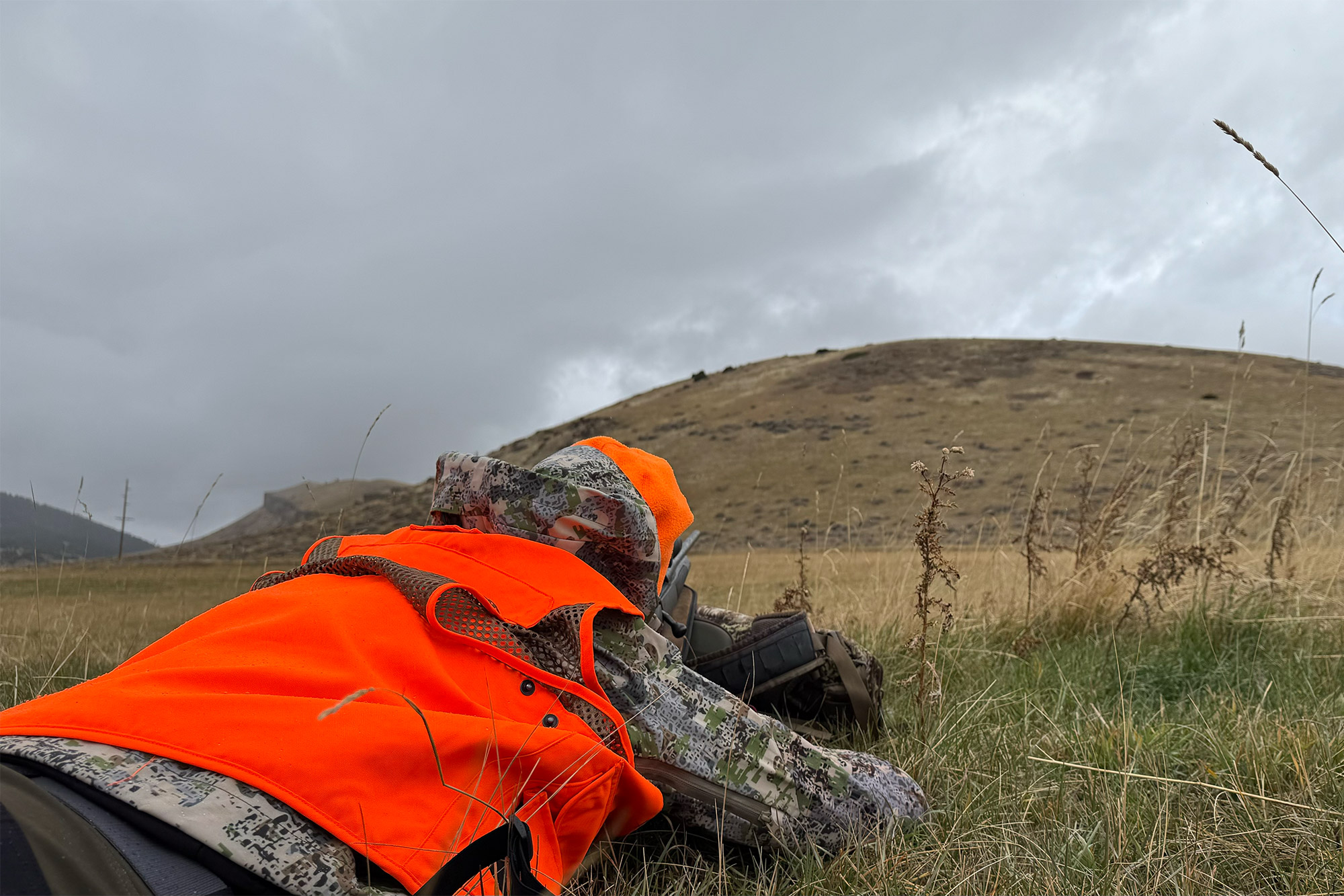 A hunter wearing a FORLOH AllClima jacket while lying in a prone position in wet grass.
