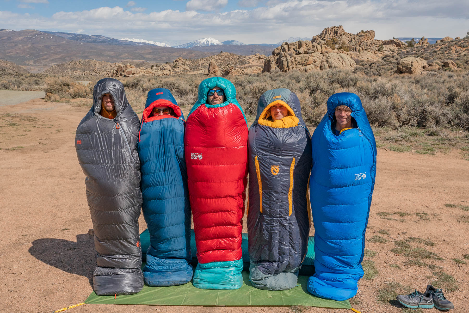 Five hikers standing outdoors in different colorful sleeping bags.