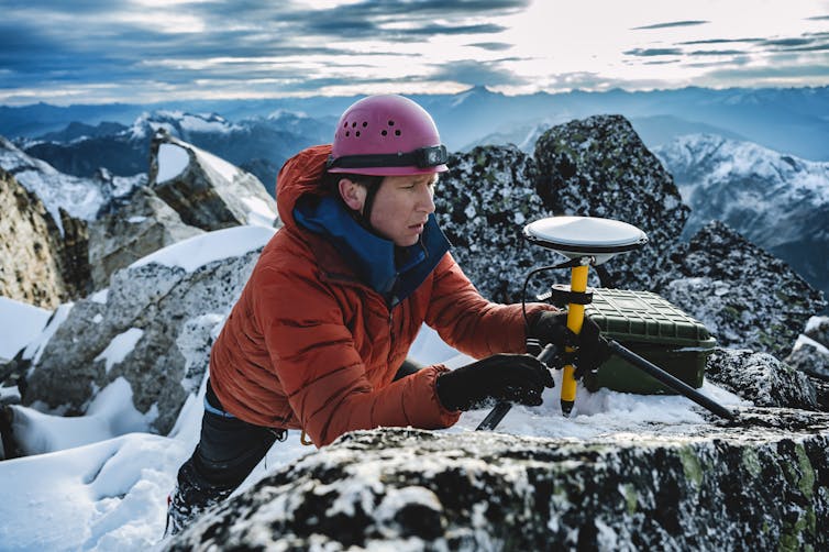 A surveyor in a red helmet and jacket carefully adjusts a measurement device on a rocky mountain top.