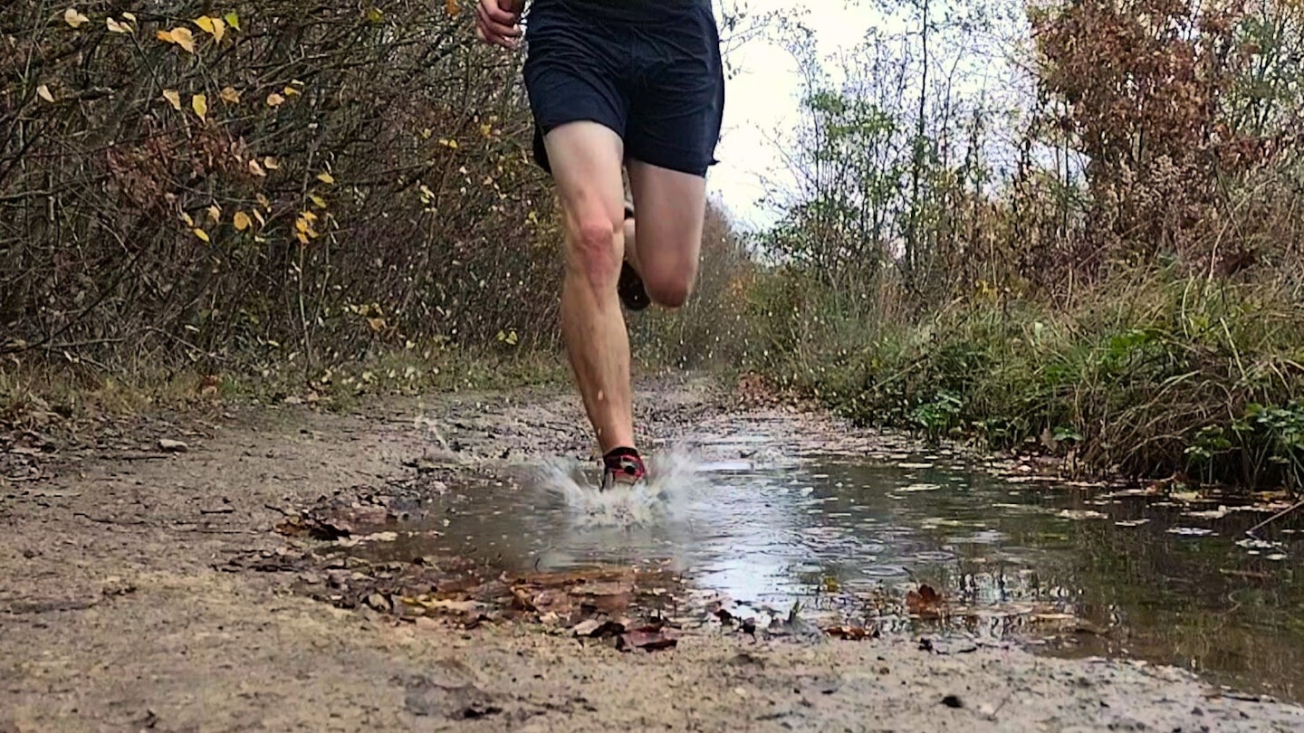 A trail runner's foot splashing through a deep puddle in a waterproof running shoe.