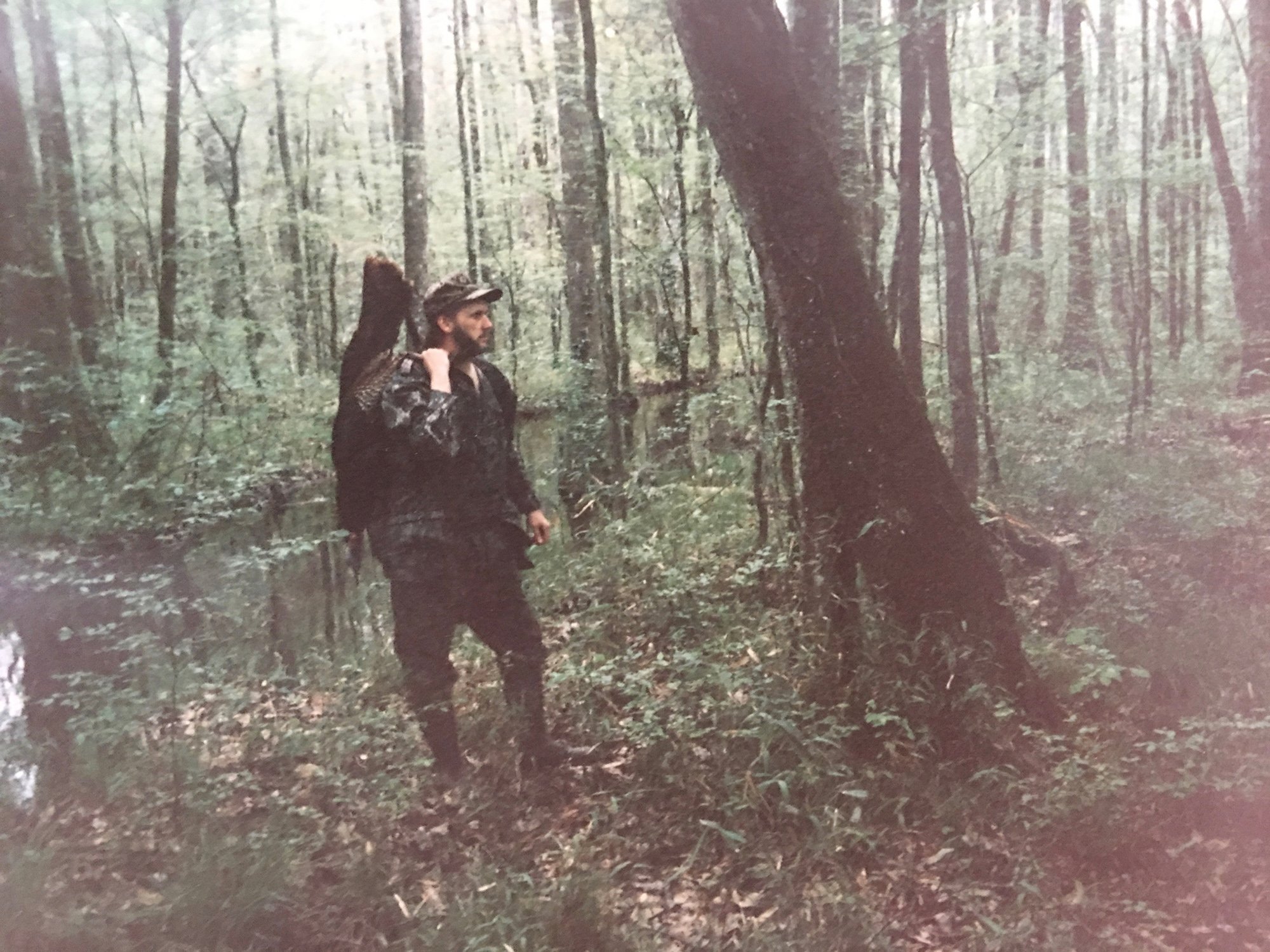 A hunter in camouflage gear posing with a harvested wild turkey in a wooded area.