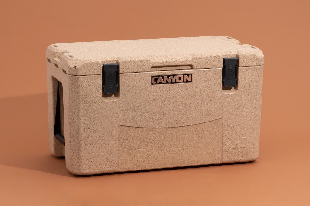 A tan Canyon Outfitter 55 cooler resting on a rocky shoreline near water