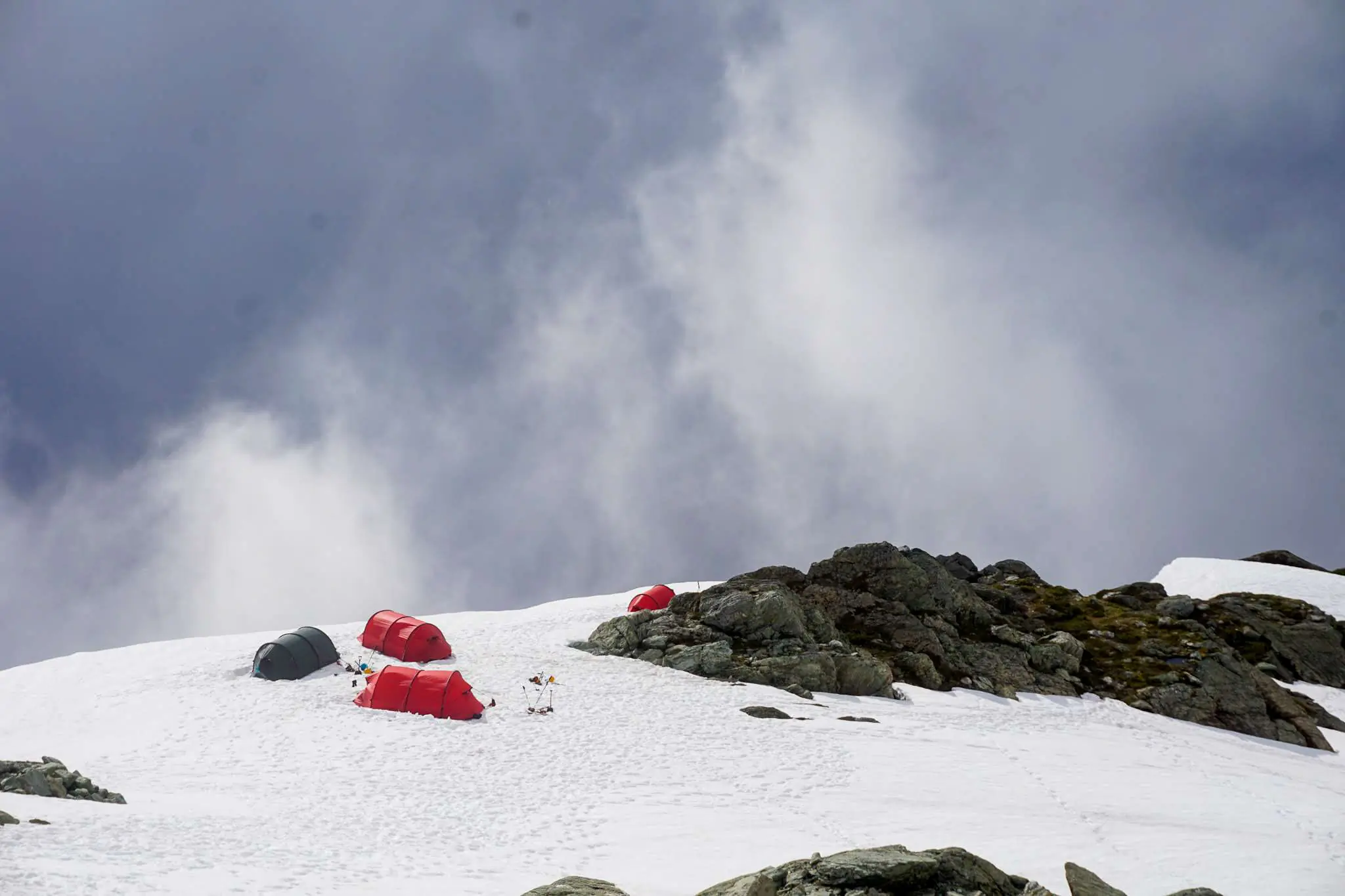 Two red Hilleberg tunnel tents pitched on a narrow snowy ridge with mountain peaks in the background.