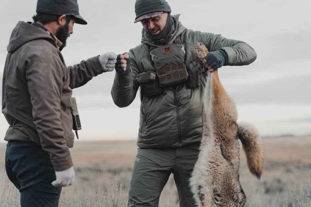 A hunter holding a coyote on the open prairie while wearing Kifaru winter gear.