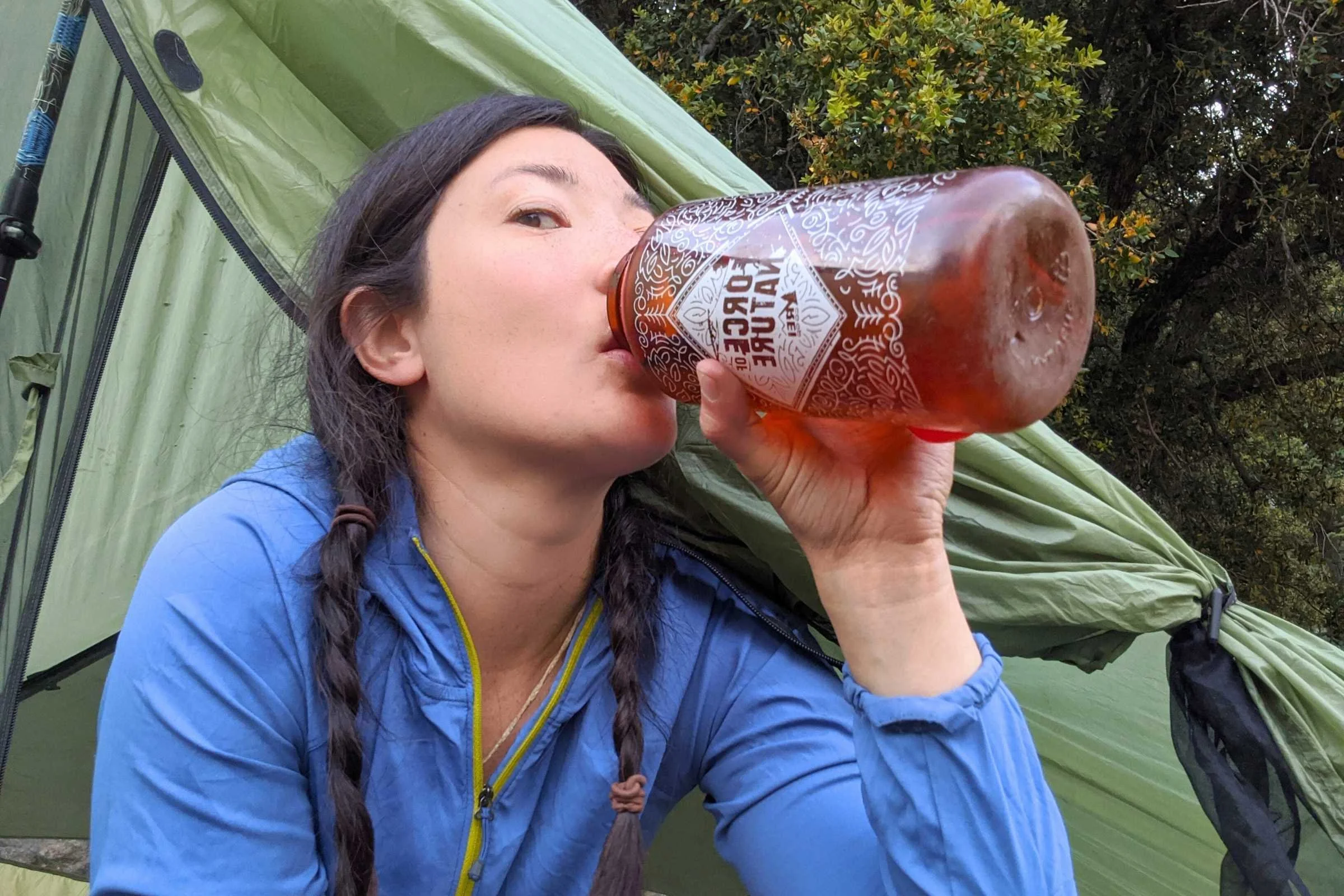 A person sitting at a mountain campsite drinking from a wide-mouth Nalgene bottle.
