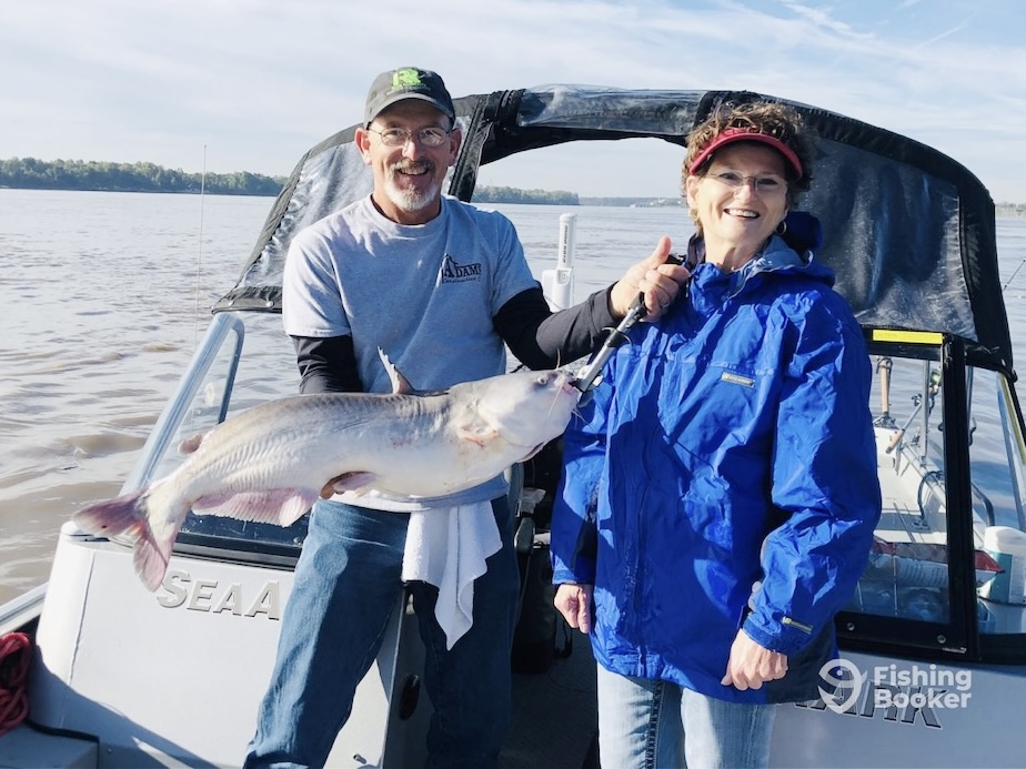 Two anglers smiling while holding a large blue catfish on a fishing boat.