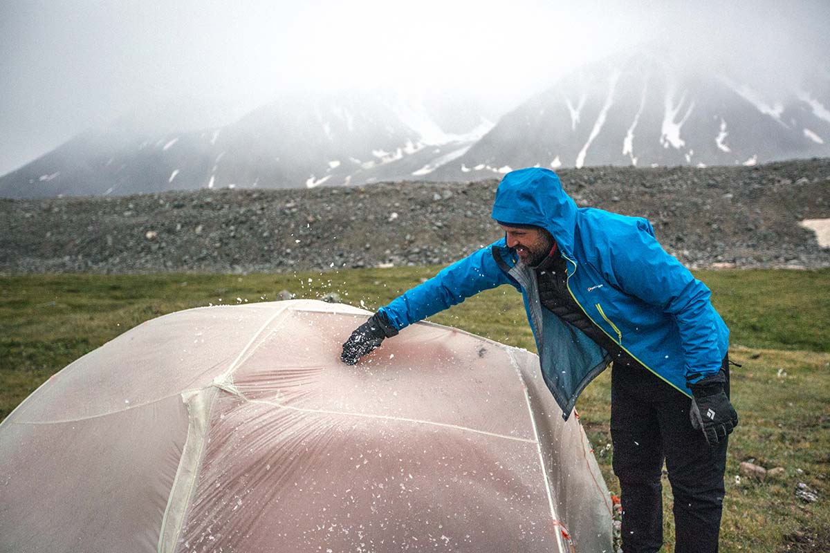 A man shaking water droplets off a grey tent fly in rainy conditions.