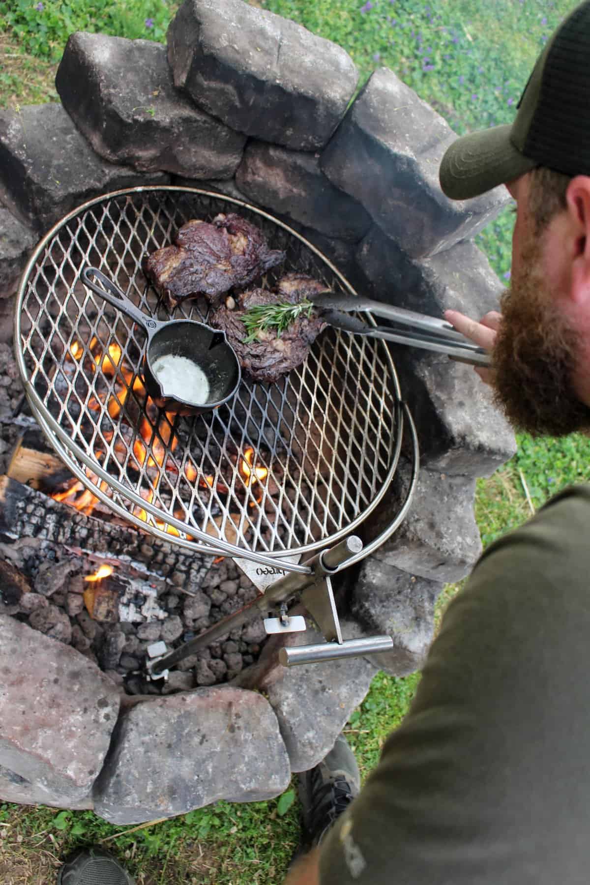 Steaks and bone marrow grilling over an open fire pit in a wilderness setting.