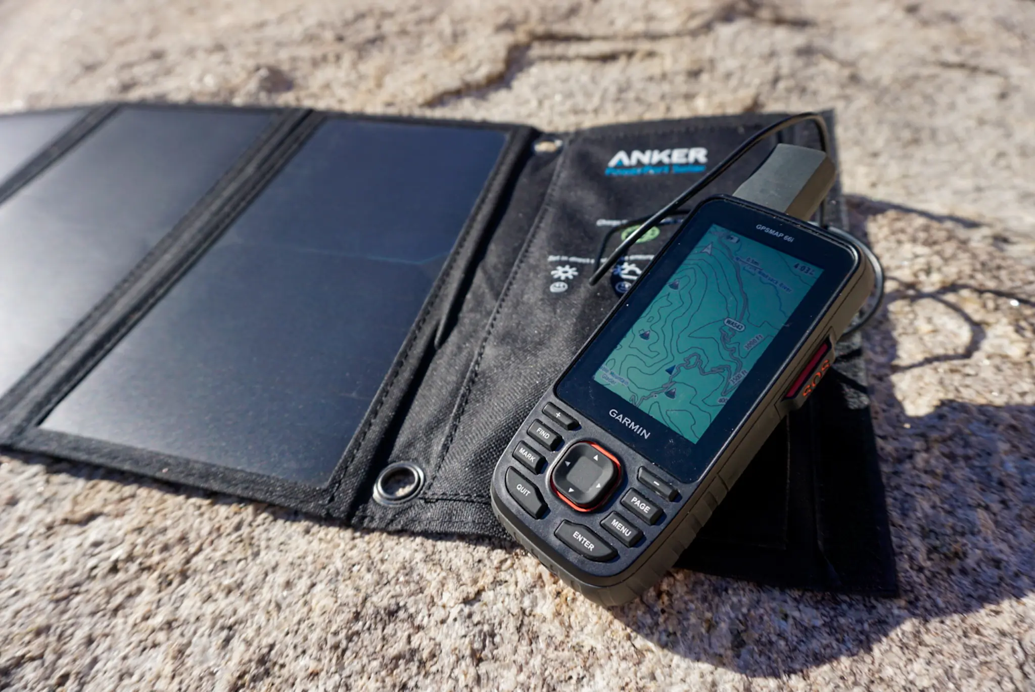 A satellite messenger connected to a portable solar panel for charging on a rock.