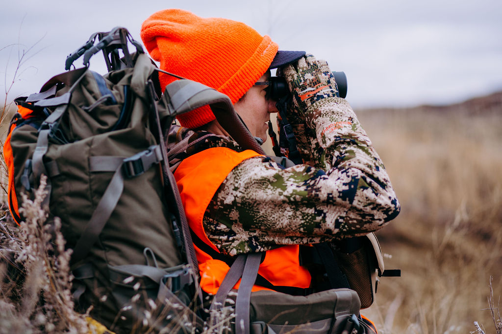 A woman in camouflage hunting gear sitting outdoors in a natural setting.