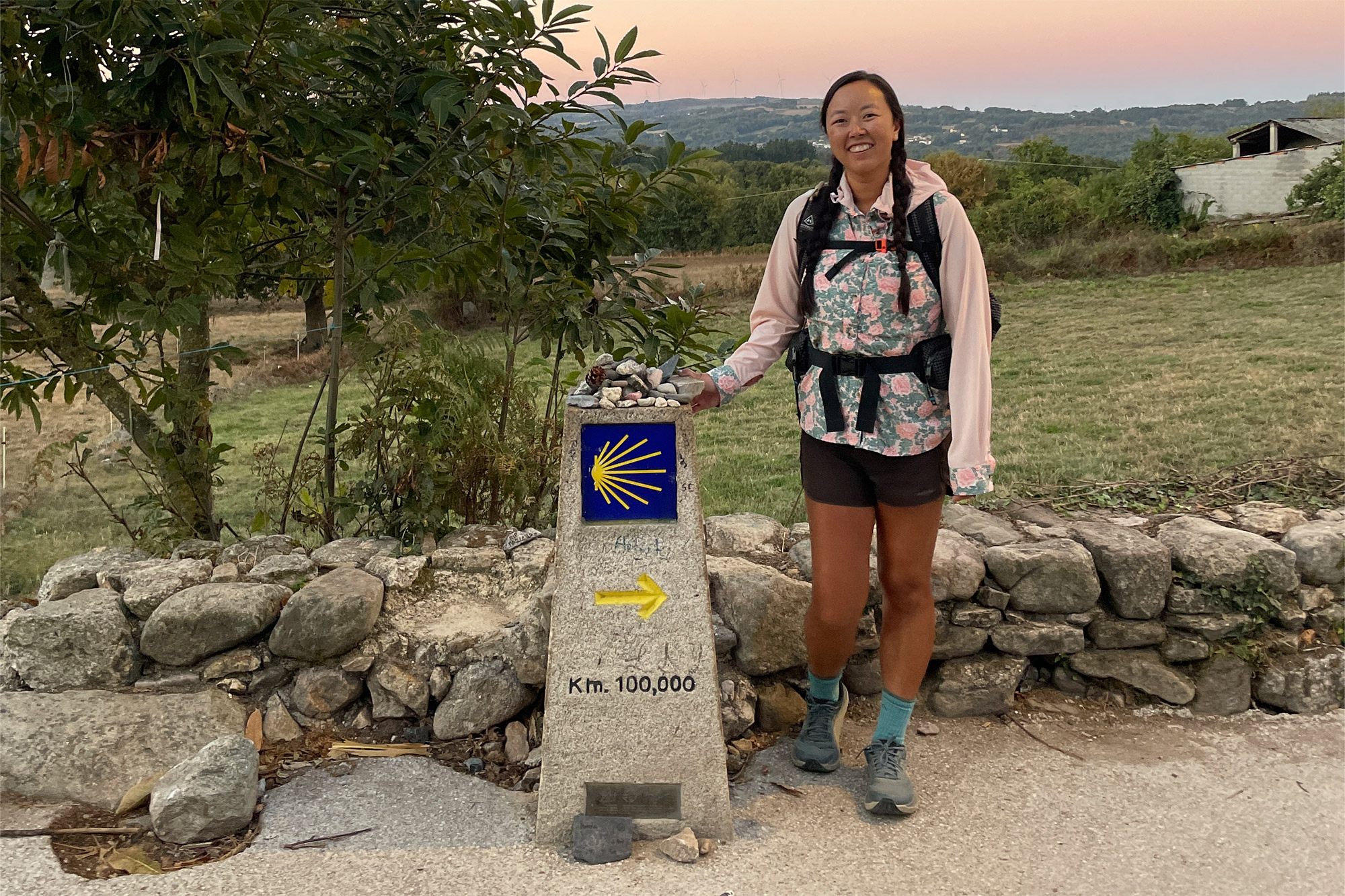 A pilgrim standing next to a stone marker carved with '100 km' on a forest path.