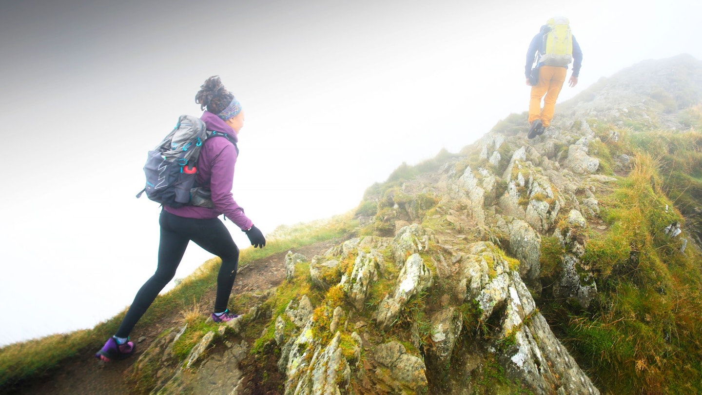 Two hikers ascending a grassy hill in overcast, rainy weather wearing waterproof gear.