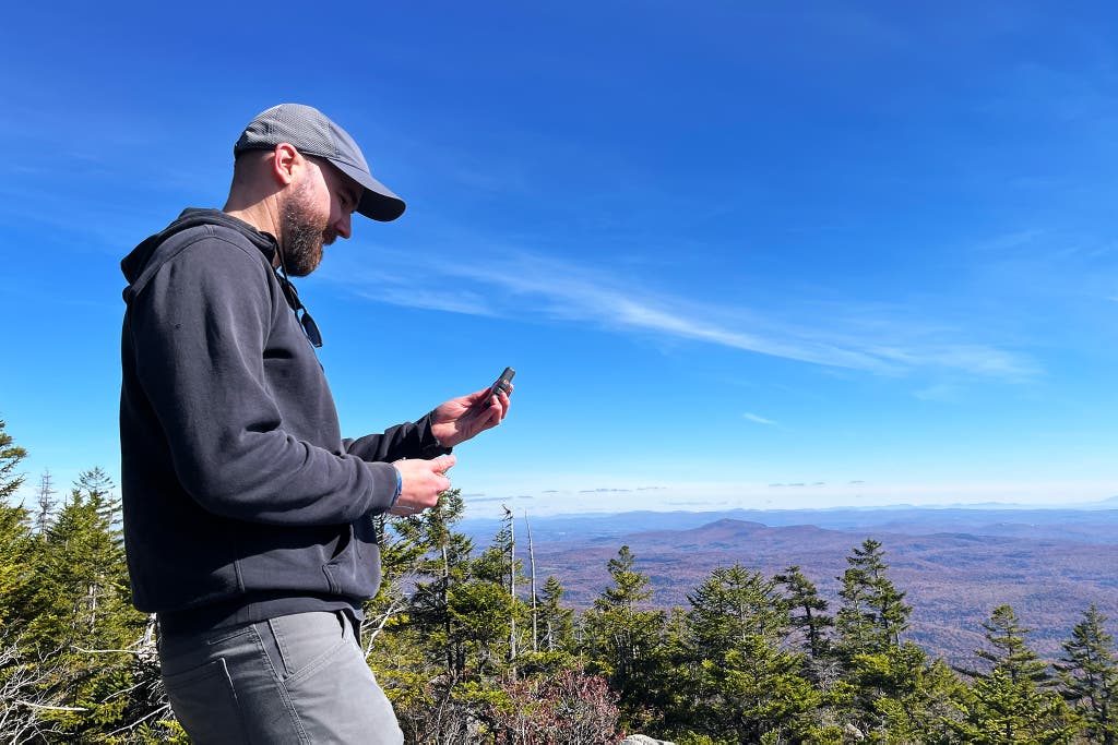 An individual holding a satellite communication device while standing in a dense evergreen forest.