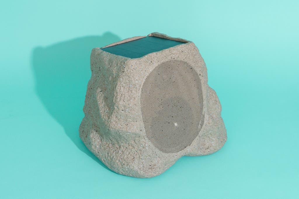 A Bluetooth speaker shaped and textured like a natural grey rock sitting in a garden bed.