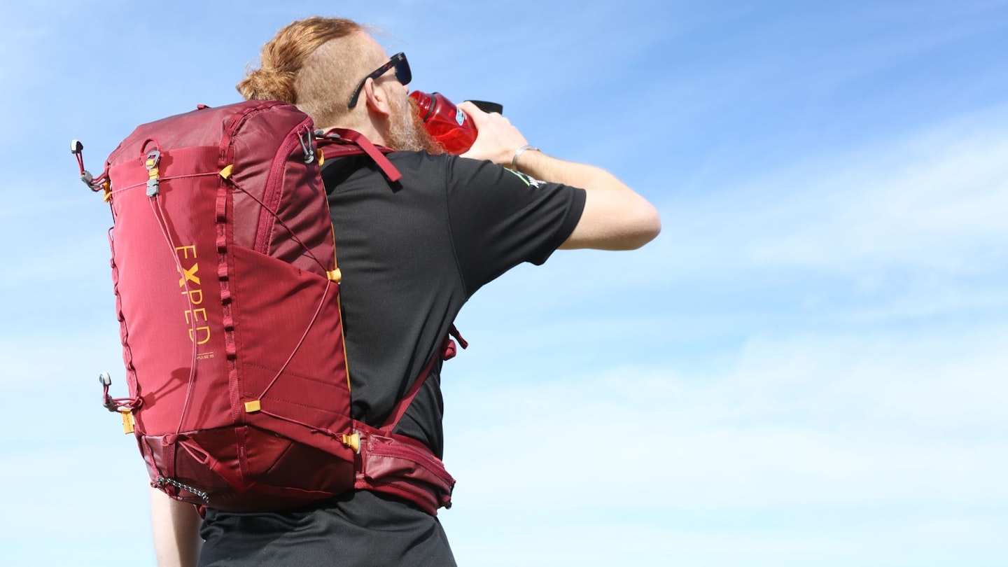 A hiker wearing a red daypack walking on a mountain trail with scenic views.