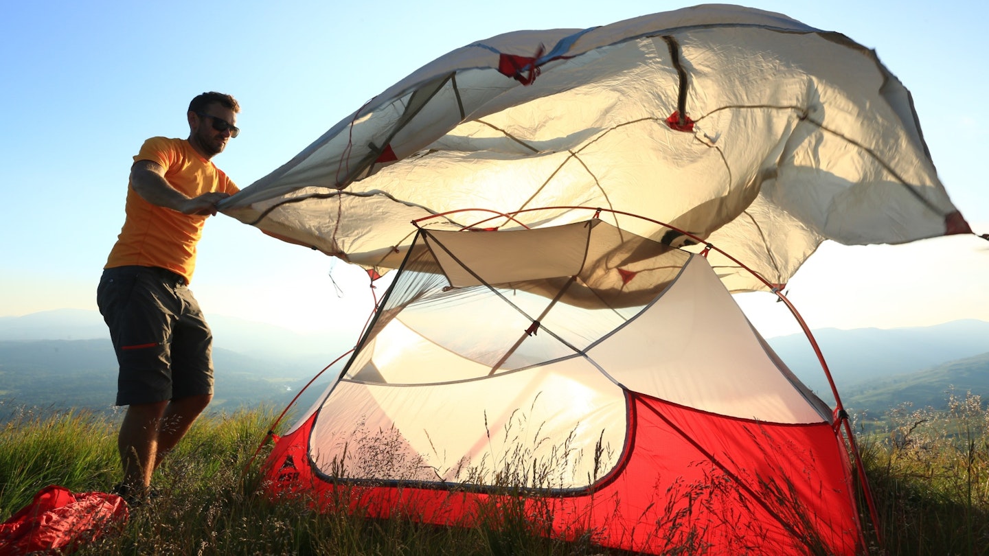 A camper placing a full-coverage flysheet over a tent frame.