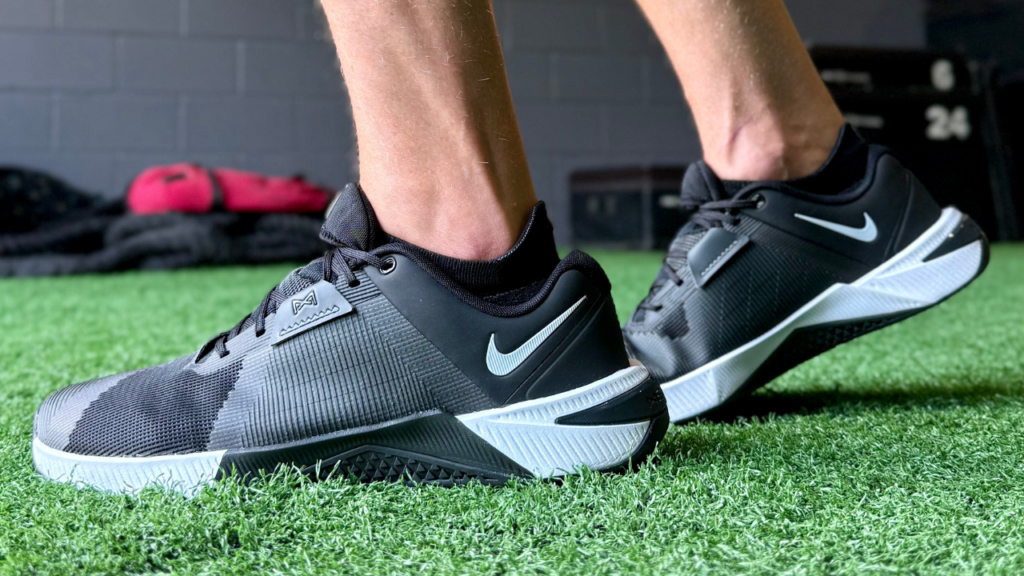 A pair of Nike Metcon 10 shoes being worn on a green turf gym floor.
